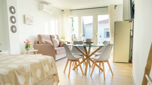 A modern bedroom with a bed, a glass dining table with chairs, a sofa, and a kitchen area. - Costa del Sol Villas - Bedroom with Private Bathroom 1 (Benajarafe)