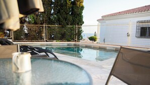 A swimming pool with a glass table and chairs in the foreground, a fenced area with trees and a building in the background. - Costa del Sol Villas - Bedroom with Private Bathroom 1 (Benajarafe)