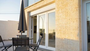 Een terras buiten met een tafel, stoelen, een grote parasol en een glazen schuifdeur die naar een overdekt gedeelte leidt.