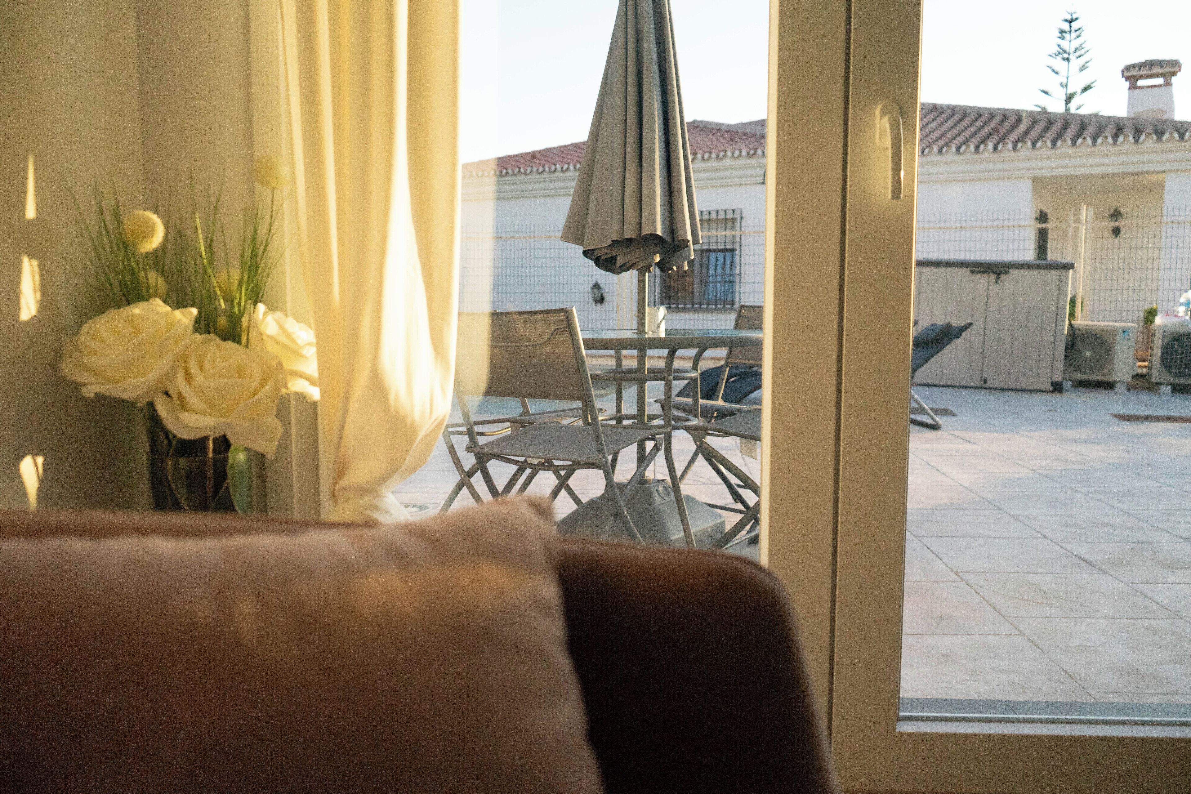 Blick aus einem Fenster mit Tisch und Stühlen davor, ein weißes Gebäude im Hintergrund und ein Blumenstrauß auf der Fensterbank.