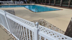 Ein Swimmingpool mit weißem Zaun und Kiesbett, eingebettet in einen Ferienort mit Palmen und Gebäuden im Hintergrund.