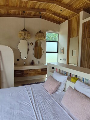 A modern bedroom with a wooden ceiling, a large bed with white and pink bedding, a vanity with a mirror, and a window with a view of greenery. - House with views of the jungle and the ocean (Santa teresa)
