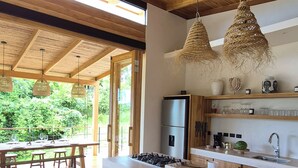 Una cocina moderna con vigas de madera, una isla grande, lámparas colgantes de mimbre y vistas a la vegetación exterior.