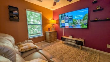 A living room with a leather couch, a flat-screen TV, a ceiling fan, a window with blinds, and a patterned rug.