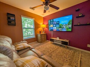 A living room with a leather couch, a flat-screen TV, a ceiling fan, a window with blinds, and a patterned rug. - 4BR Home with calif king bed Sleeps 10, Gym near Airport (Atlanta)