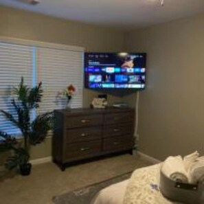 Une pièce avec un téléviseur fixé au mur, une commode en bois foncé, une plante en pot et un lit recouvert d’une couvre-lit à motifs.