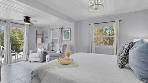 A bedroom with a large bed, a chair, a table, and a ceiling fan. There are windows with curtains, a ladder, and a view of trees outside.