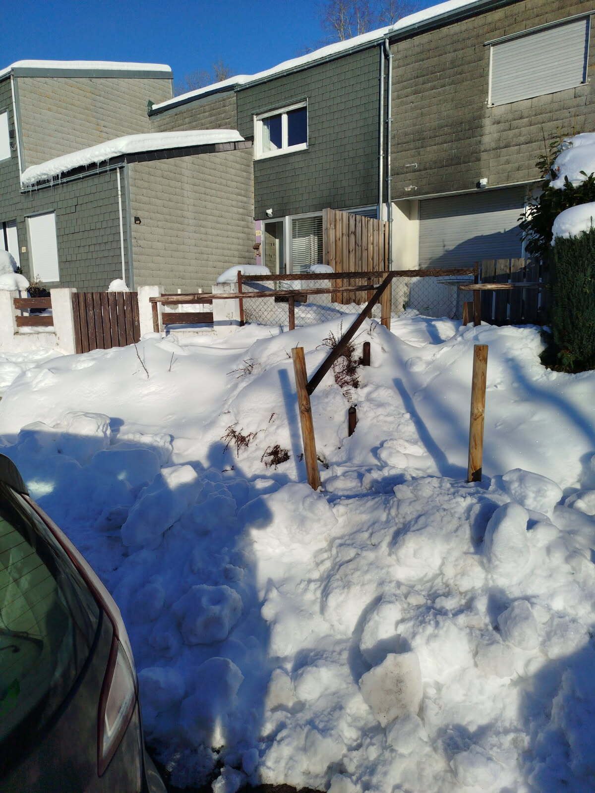 A residential area with snow-covered ground, a wooden fence, and a car partially visible.