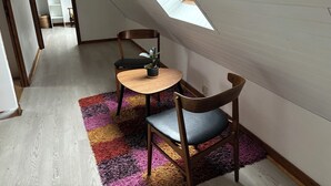 Een kleine, gezellige kamer met schuine wanden en dakraam, met een houten tafel, twee stoelen en een kleurrijk vloerkleed.