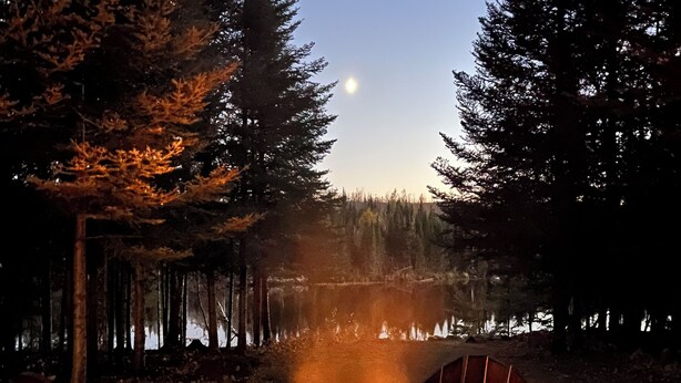 A campfire with two chairs and a table in a forested area with a lake in the background.