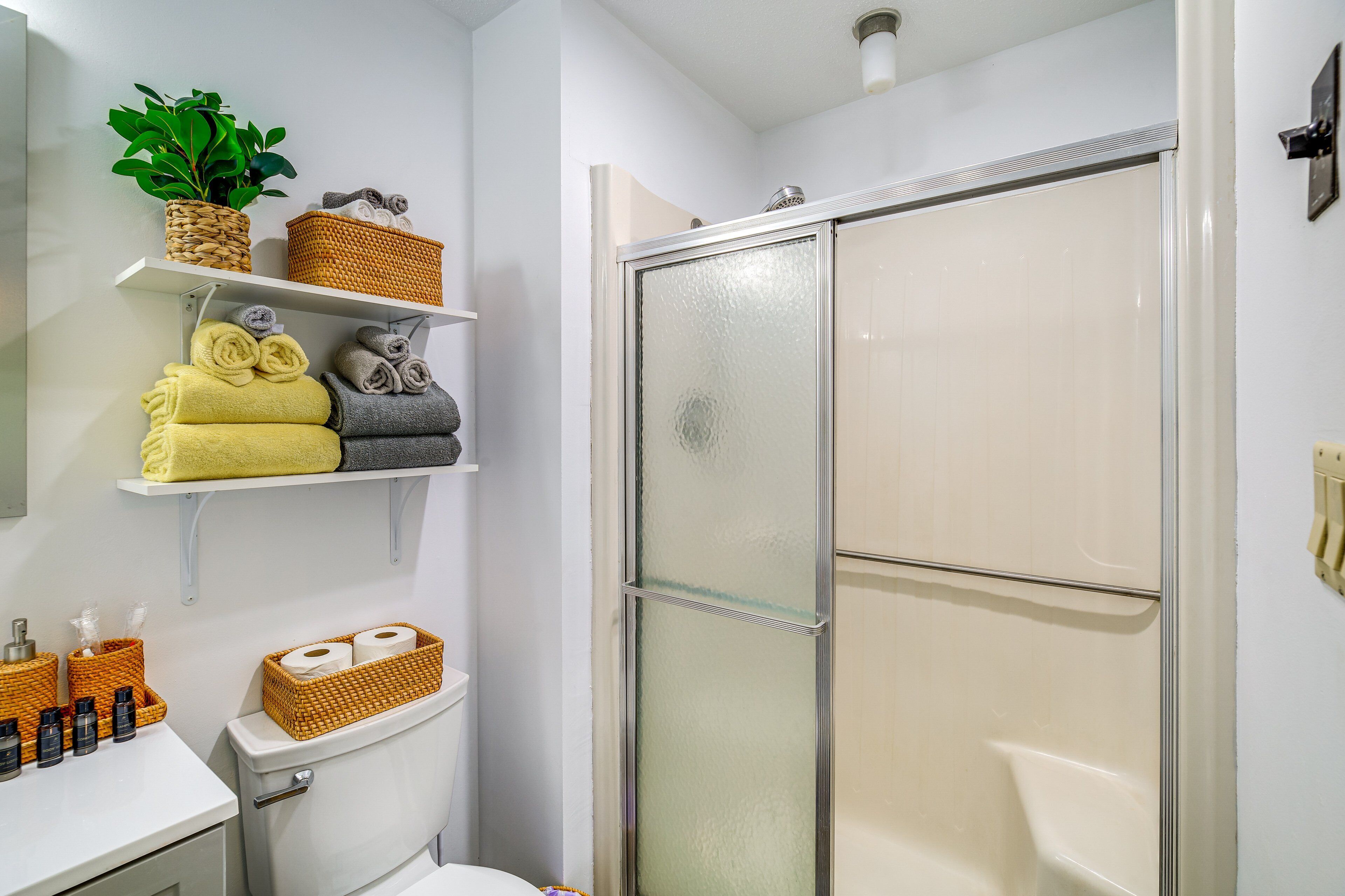 Casa de banho com sanita, chuveiro com portas de vidro jateado, prateleiras com toalhas e cestos, e uma planta em vaso.