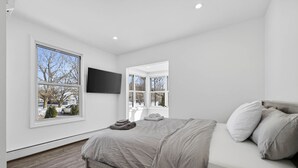 A bedroom with a bed, a TV mounted on the wall, and two windows.