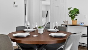 A dining area with a round wooden table set for six, surrounded by chairs. There is a potted plant on the table, and a small table with a plant and a water bottle in the background.