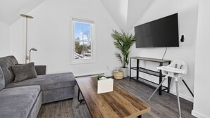 Una sala de estar moderna con un sofá seccional gris, una mesa de centro de madera, un televisor montado en la pared, un pequeño escritorio con una silla y una planta en maceta.