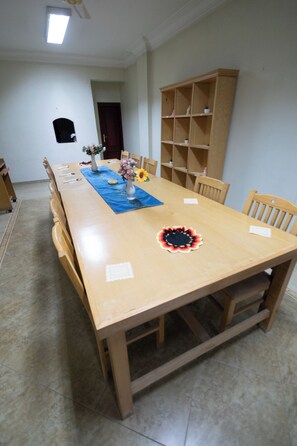 Ein Esszimmer mit einem langen Holztisch, Stühlen, einer blauen Tischläufer, einer Vase mit Blumen und einem Bücherregal.