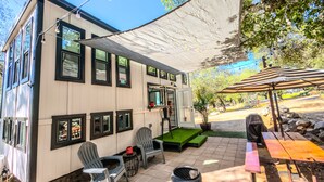 Ein zweistöckiges Gebäude mit einer Terrasse, die Sitzgelegenheiten im Freien, einen Grill und einen beschatteten Bereich mit einem gestreiften Sonnenschirm bietet.