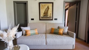 Um sofá em tons neutros com duas almofadas, um vaso com plantas secas e um quadro de um veleiro na parede.