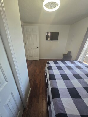 Um quarto com colcha xadrez, ventilador de teto, televisão de tela plana na parede e porta para outro cômodo.
