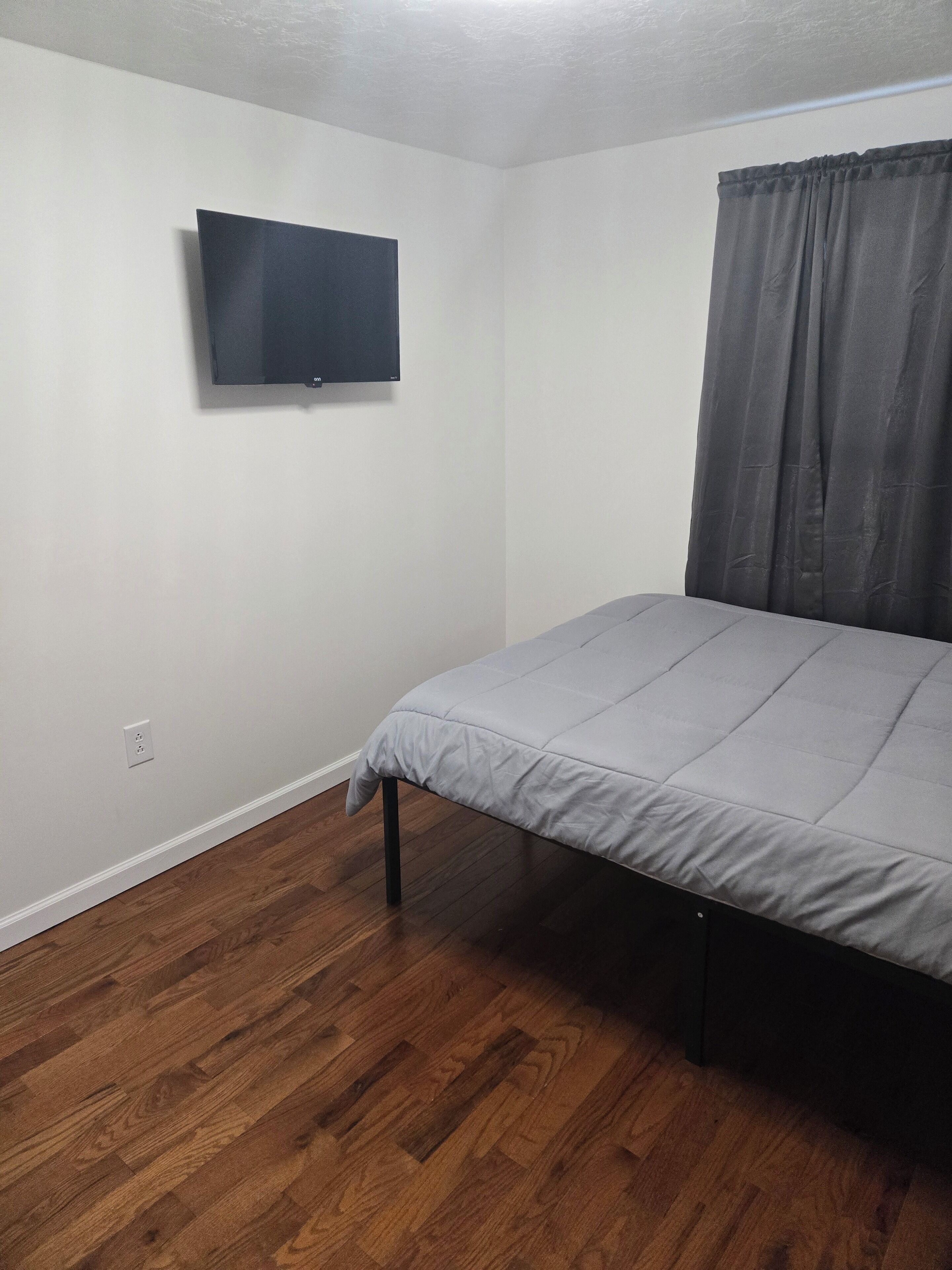 Un dormitorio con una cama, televisión montada en la pared y una ventana con cortinas.