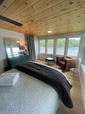 A bedroom with a bed, a dresser, a chair, and a view of a lake through large windows.