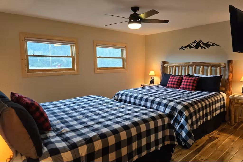 Ein Zimmer mit zwei Betten, jeweils mit karierten Laken, einem Deckenventilator, zwei Fenstern und einem an der Wand befestigten Fernseher.