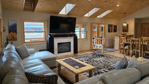 A spacious living room with a fireplace, wooden ceiling, and large windows. - Trout Hunter Cabin- South Fork (South Fork)