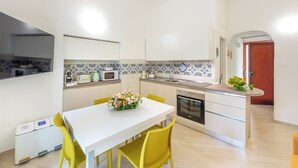 Una cocina moderna con armarios blancos, un microondas y un fregadero. Hay una mesa de comedor con sillas amarillas y un centro de mesa floral.