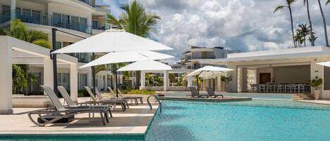 Un área de piscina con sillones de descanso y sombrillas, rodeada de edificios modernos y palmeras.