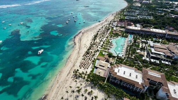 Vista aérea de un complejo turístico costero con playa, piscinas y un puerto deportivo.