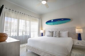 Quarto de hotel com cama, duas mesas de cabeceira, ventoinha no teto, janela grande com cortinas translúcidas e pintura na parede retratando uma praia.