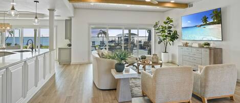 A spacious living area with a view of the sea, a dining table with chairs, and a television on the wall.