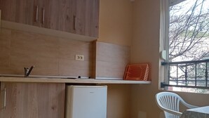 A compact kitchen with wooden cabinets, a white refrigerator, a small trash bin, and a window with a view of trees. - Classic Triple Room with Mountain View, Loggia and Shared Bathroom (Sutomore)