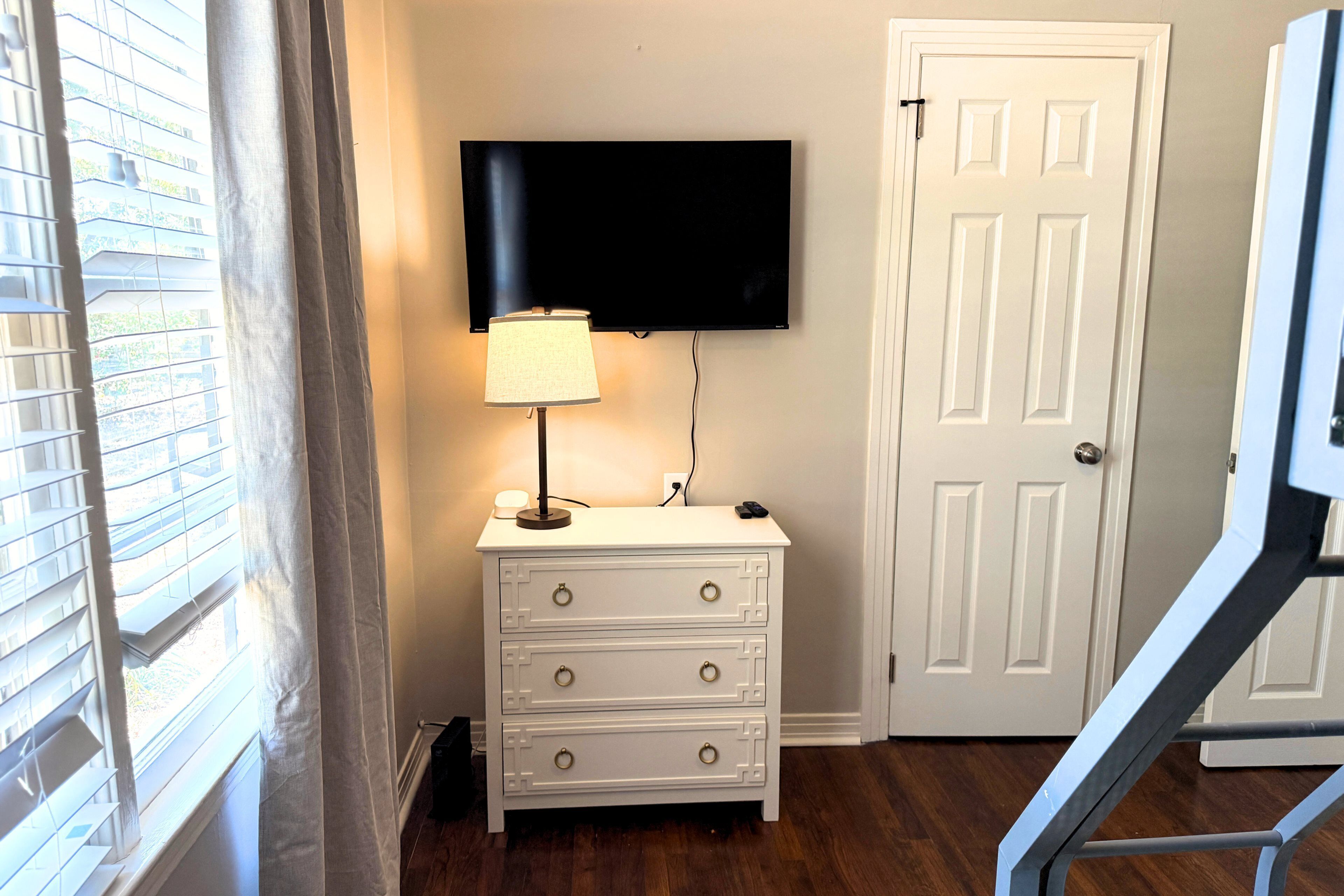 A room with a white dresser, a mounted TV, a lamp, and a door.
