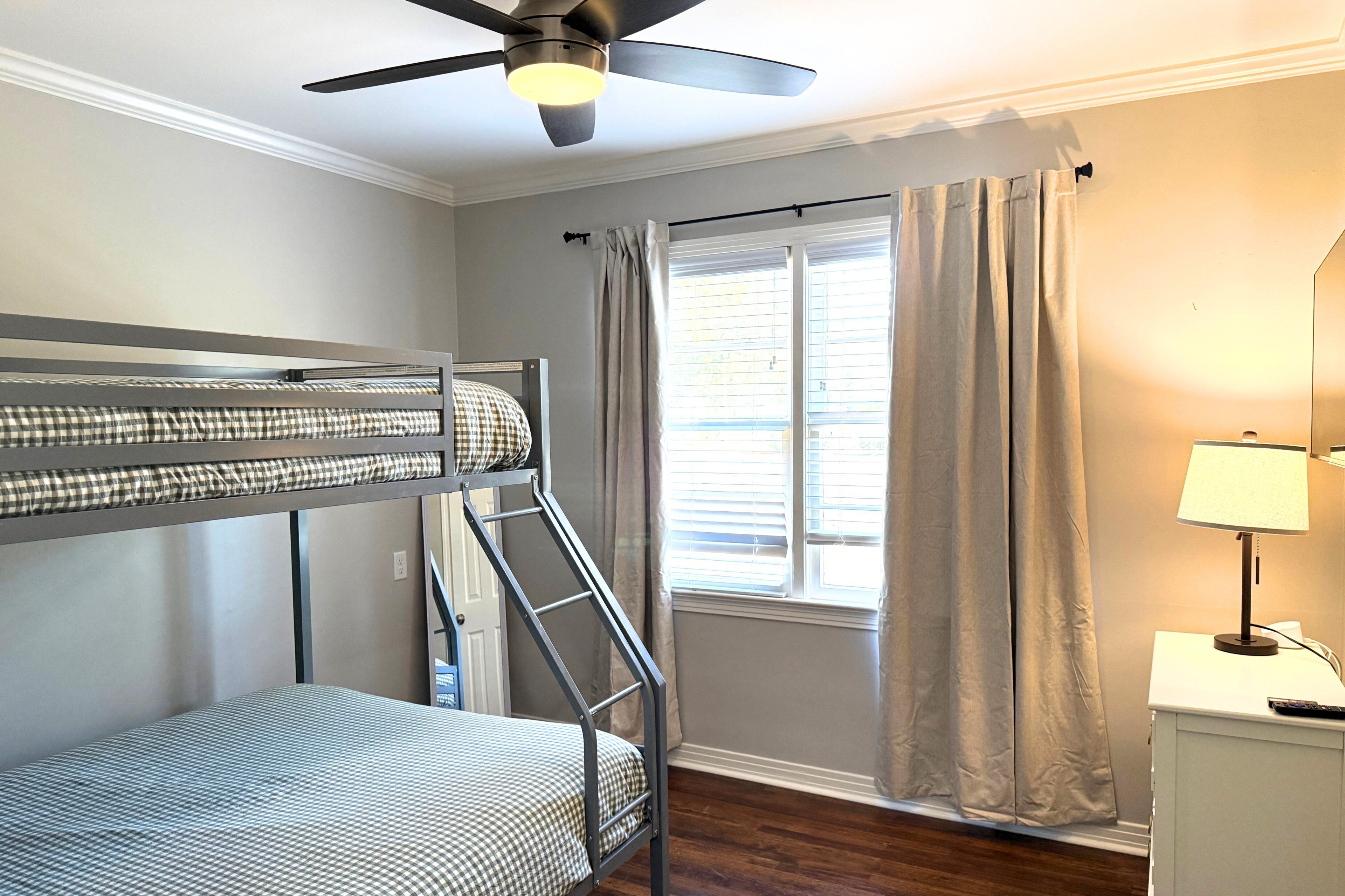 A bedroom with a bunk bed, a ceiling fan, a window with curtains, and a bedside table with a lamp.