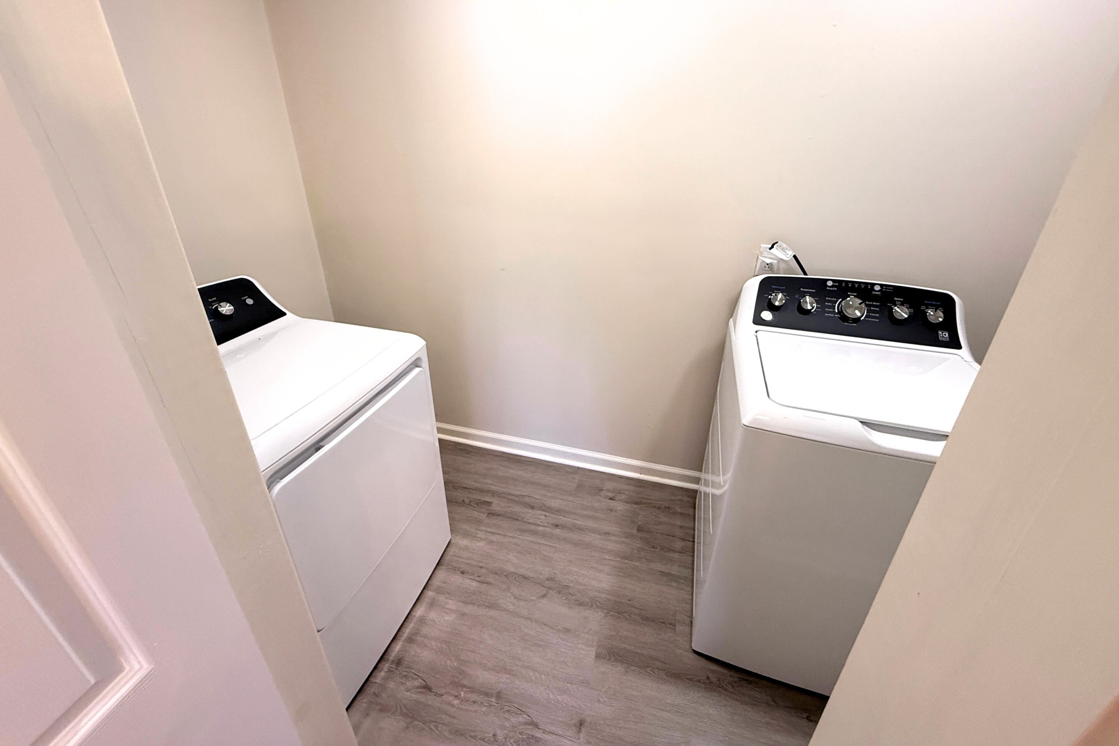 A laundry room with two white front-loading washing machines and a dryer, wooden flooring, and light-colored walls.