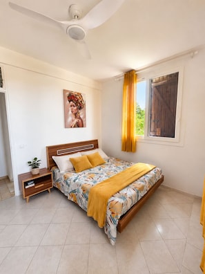 Une chambre à coucher avec un lit, une table de chevet, un ventilateur de plafond, une fenêtre avec des rideaux et un tableau au mur.