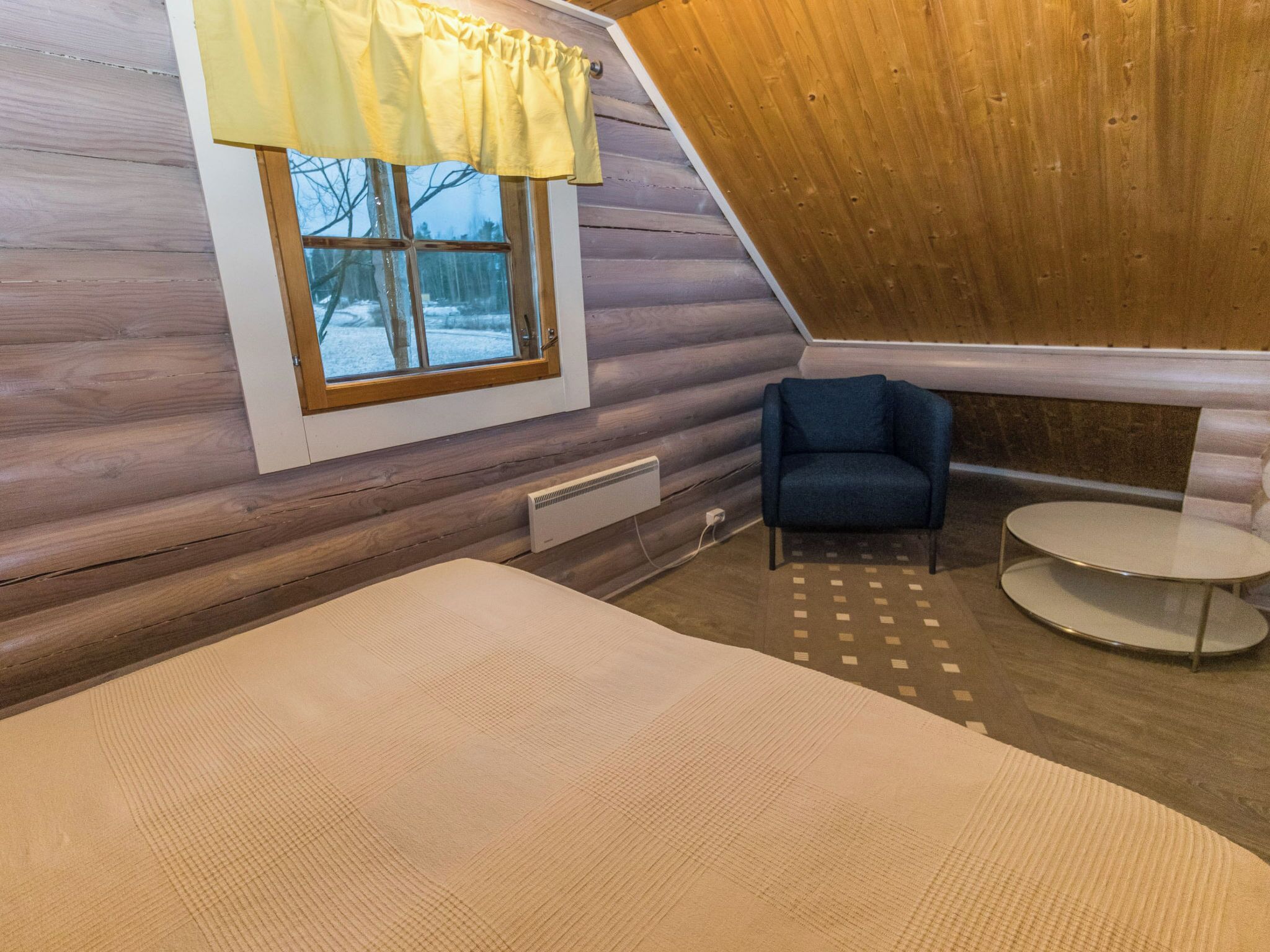 Una habitación acogedora con techo de madera inclinado, una ventana con cortina amarilla, una silla azul y una pequeña mesa redonda.