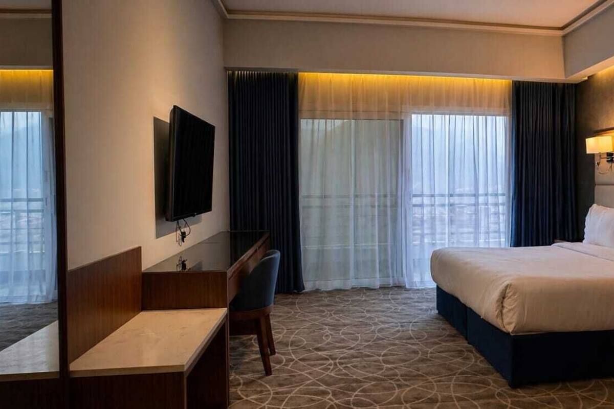 Ein Hotelzimmer mit einem Bett, einem Schreibtisch, einem Fernseher und einem großen Fenster mit Vorhängen.