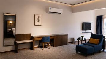 A hotel room with a desk, chair, sofa, TV, air conditioning unit, and a mirror.
