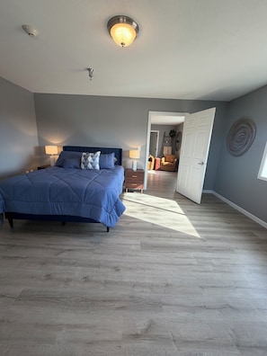 Une chambre à coucher avec un grand lit, deux tables de chevet avec des lampes, un luminaire au plafond et une porte donnant sur une autre pièce.