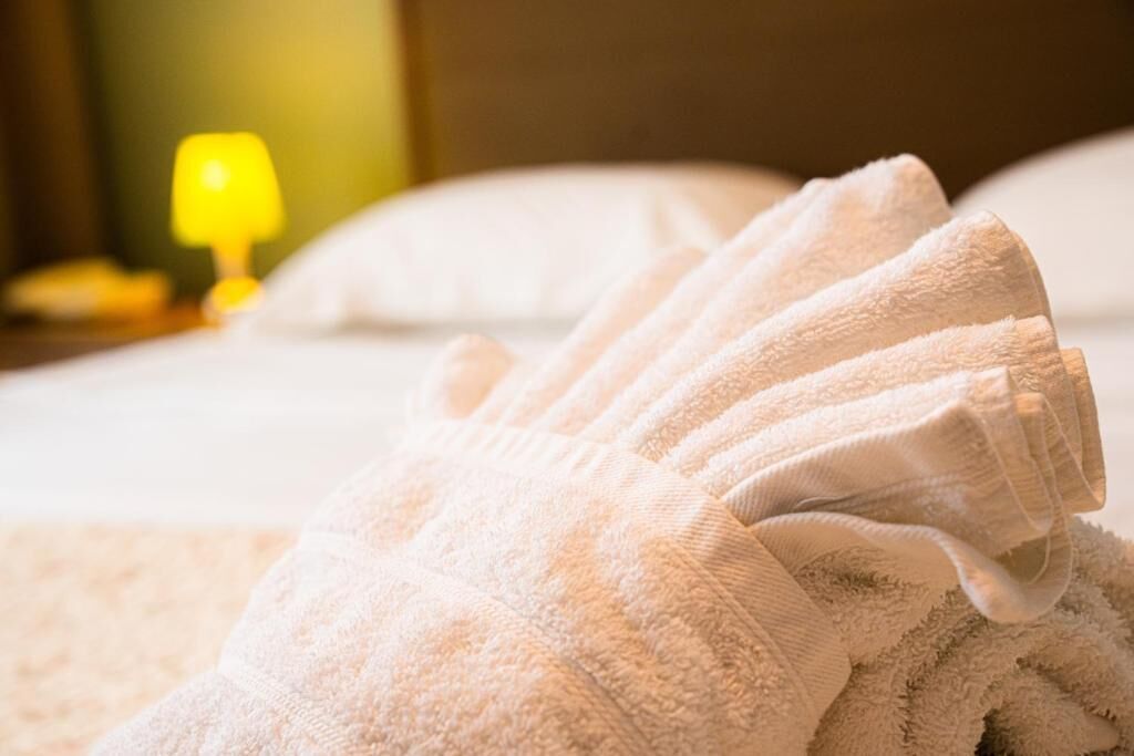 A neatly folded white towel on a bed with a visible headboard and a lamp in the background.
