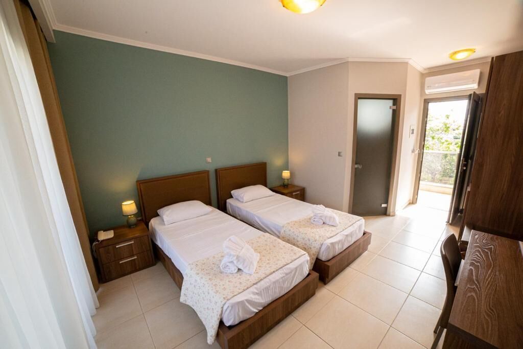 A hotel room with two beds, a wooden headboard, bedside tables with lamps, a wooden desk, and a door leading to a balcony.