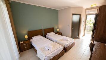 A hotel room with two beds, a wooden headboard, bedside tables with lamps, a wooden desk, and a door leading to a balcony.