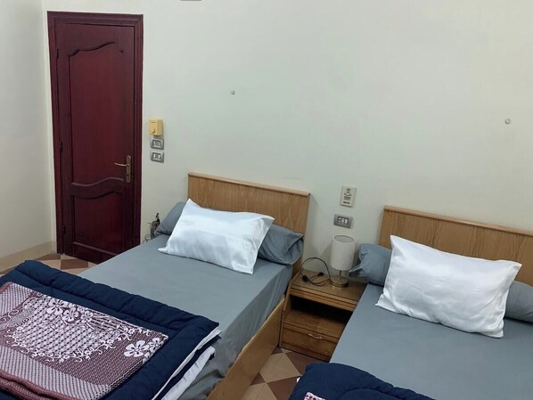 A room with two beds, each with a pillow and a nightstand. There is a wooden door and a wall socket.