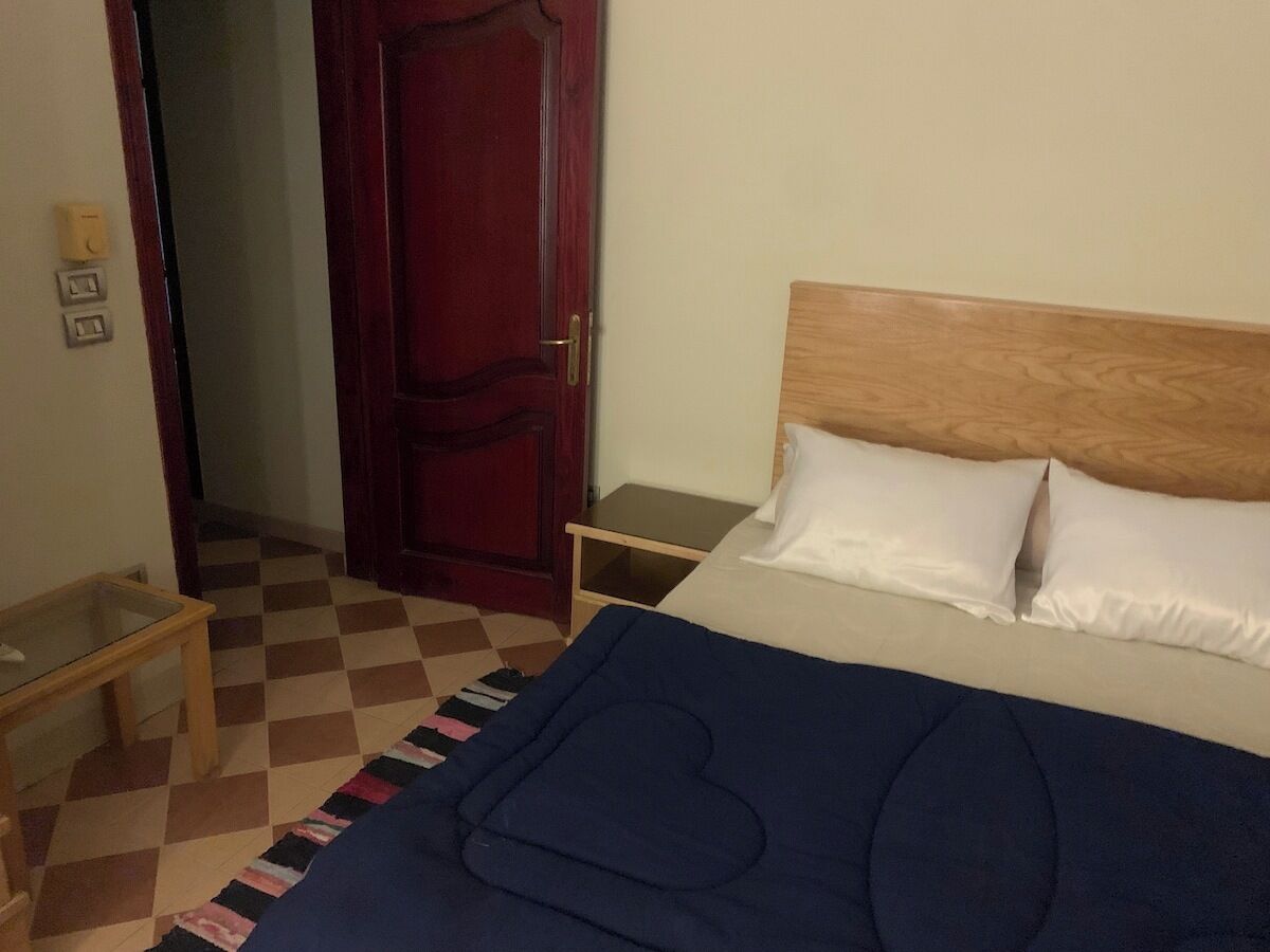 Een slaapkamer met een bed, een houten hoofdeinde, een nachtkastje en een geruit vloertje.