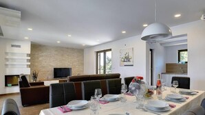 A dining area with a long table set for six, a sofa, and a television in the background. - Luxury 5 Bed, 7 Bath, Villa. Pool, Gym, Sauna, Hottub, Parking, 2 terr w/Seaview (Jursici)