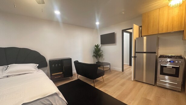Une chambre d’hôtel moderne équipée d’un lit, d’une chaise noire, d’une petite table, d’une plante en pot, d’un téléviseur et d’un coin cuisine avec un réfrigérateur et une cuisinière.