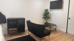 Une chambre moderne avec un lit, un canapé noir, une petite table ronde, une plante en pot et un téléviseur à écran plat fixé au mur.