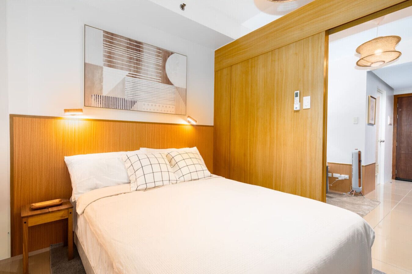 A hotel room with a bed, wooden headboard, a nightstand, a wall-mounted light, and a door leading to another room.