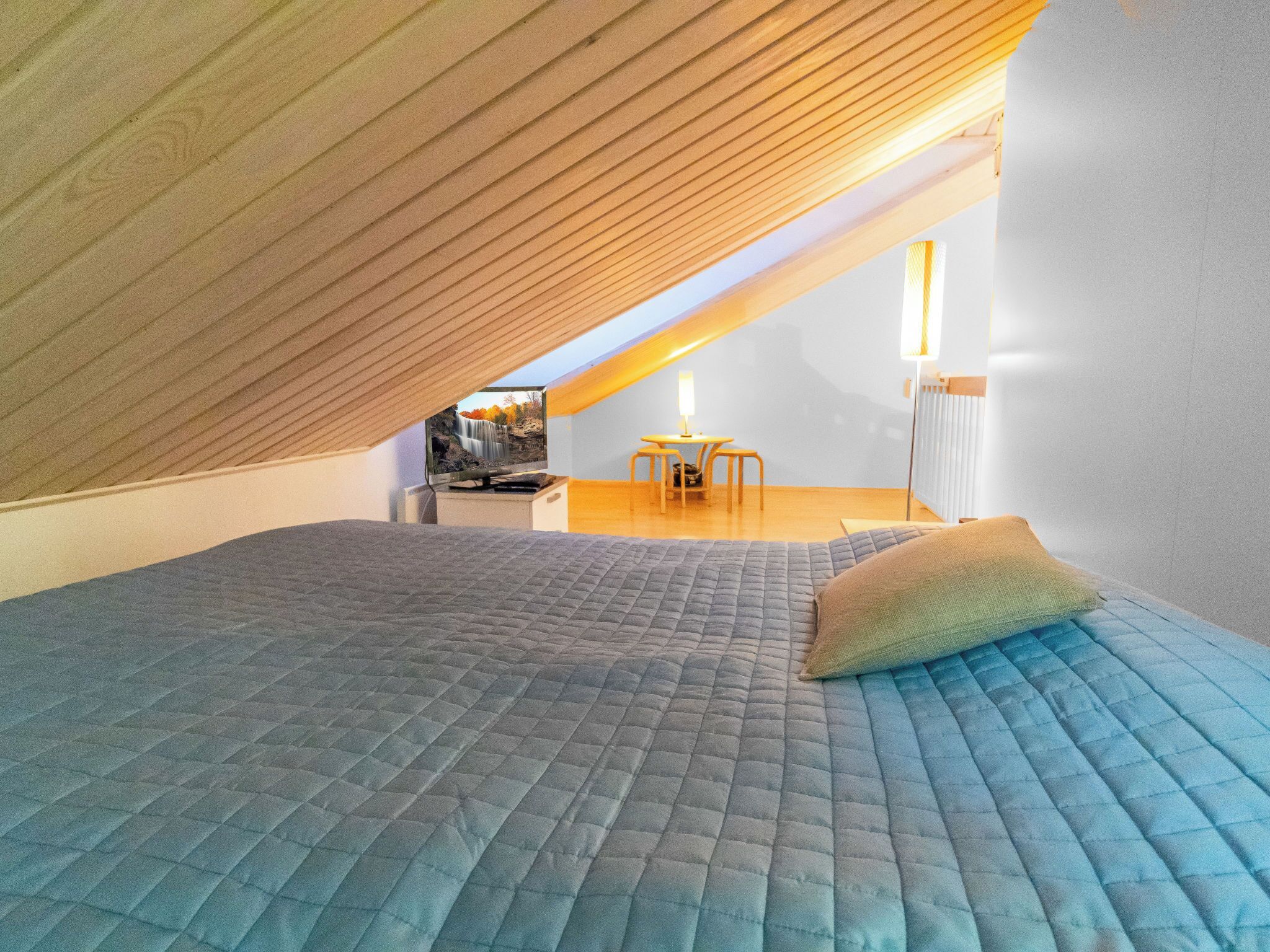 Une chambre avec un plafond en pente, un lit recouvert d’une courtepointe, une petite table avec une chaise et un téléviseur posé sur un meuble.